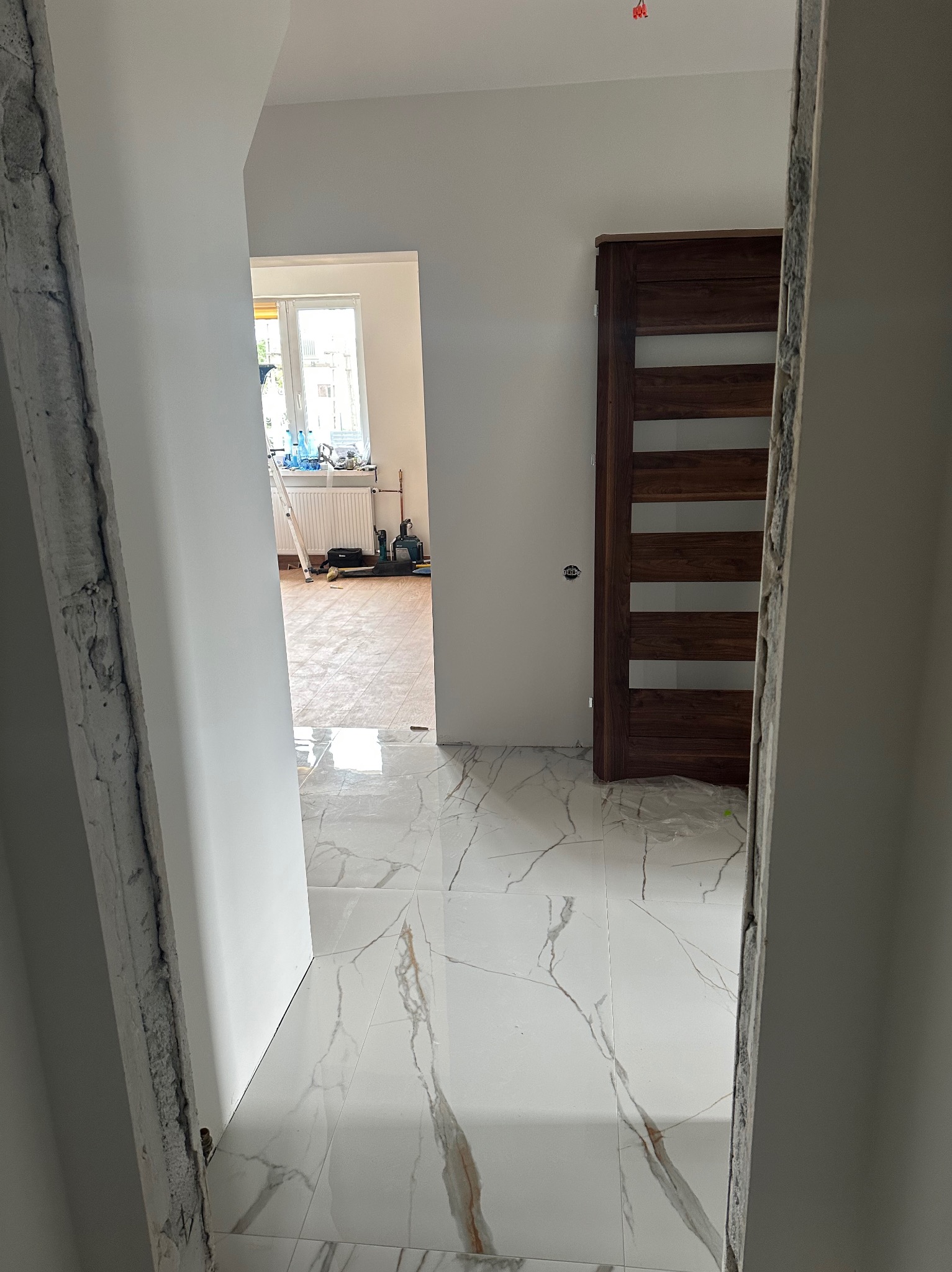 Nowoczesne wnętrze w trakcie wykańczania: białe marmurowe płytki podłogowe, drewniane drzwi z przeszkleniami i surowy betonowy fragment ściany.