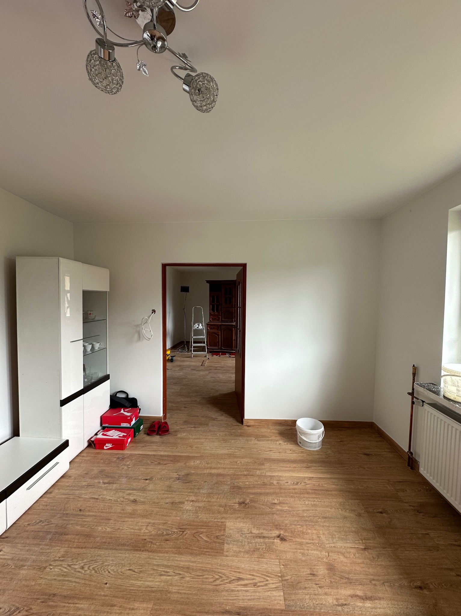 Jasne wnętrze po odświeżeniu: świeżo pomalowane ściany, panele podłogowe, biała meblościanka. W tle drabina malarska i otwarte przejście do kolejnego pomieszczenia.