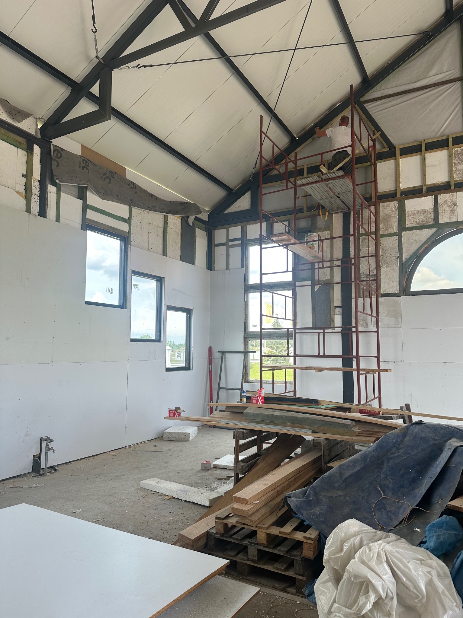 Wnętrze w trakcie wykończenia: ściany z płyt gipsowo-kartonowych, okna, rusztowanie z pracownikiem przy suficie. Widoczne materiały budowlane i narzędzia.