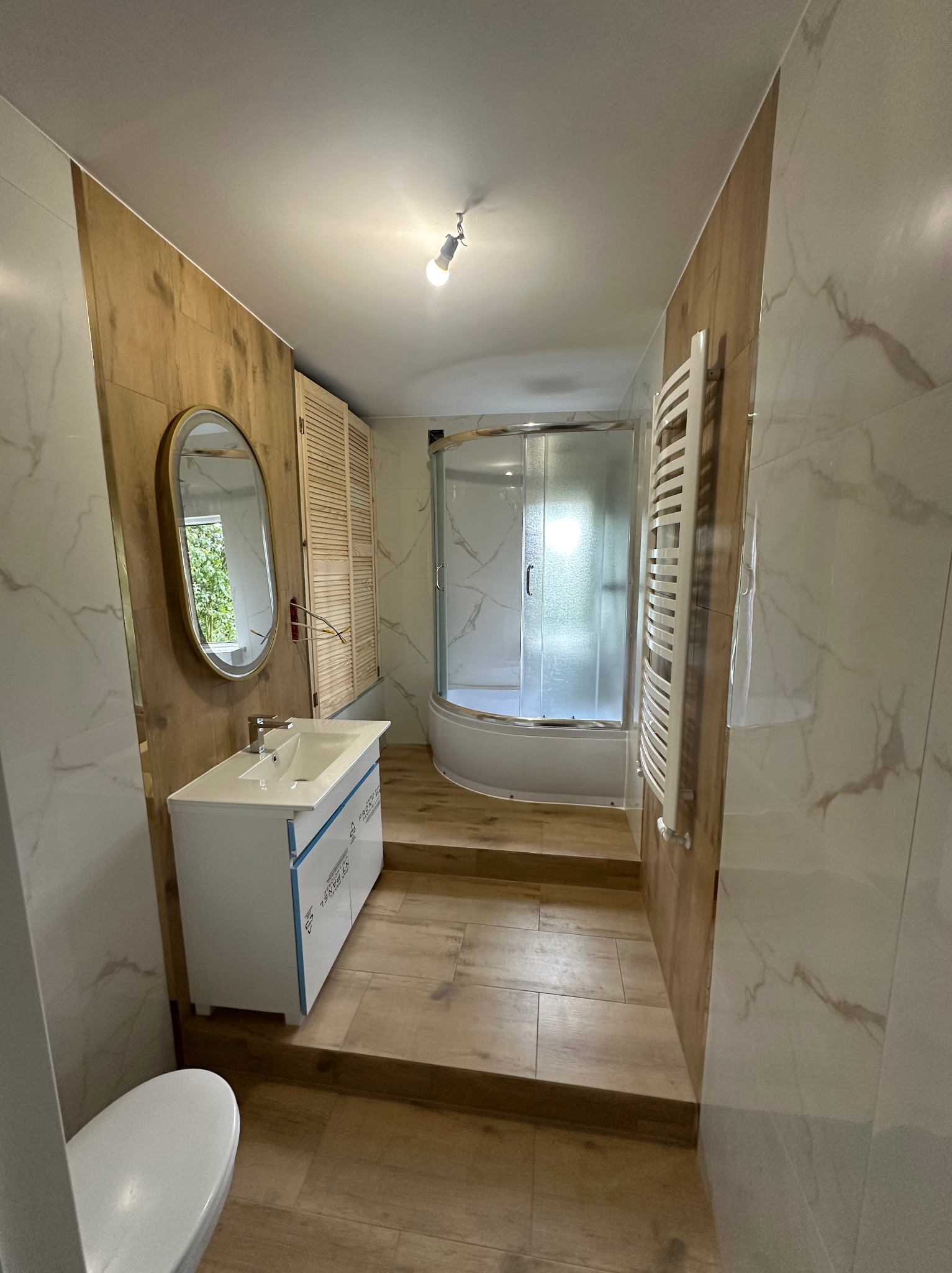 Nowoczesna łazienka z kabiną prysznicową, umywalką z szafką i lustrem w złotej ramie. Płytki imitujące drewno i marmur tworzą elegancki wystrój.