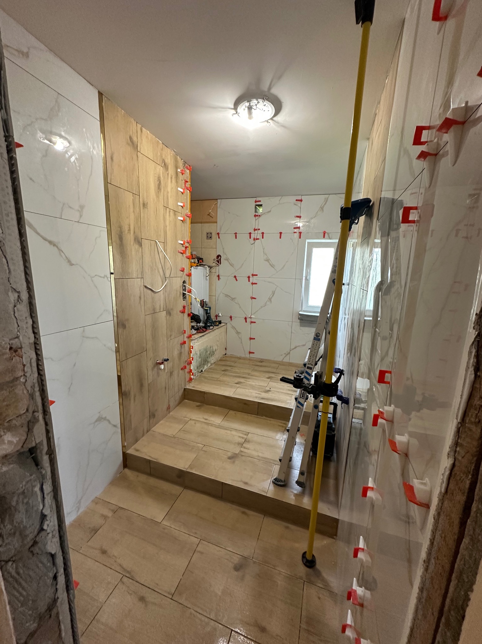 Układanie płytek imitujących drewno i marmur w łazience, widoczne poziomice i system poziomowania płytek. Prace glazurnicze w toku, z dbałością o detale.