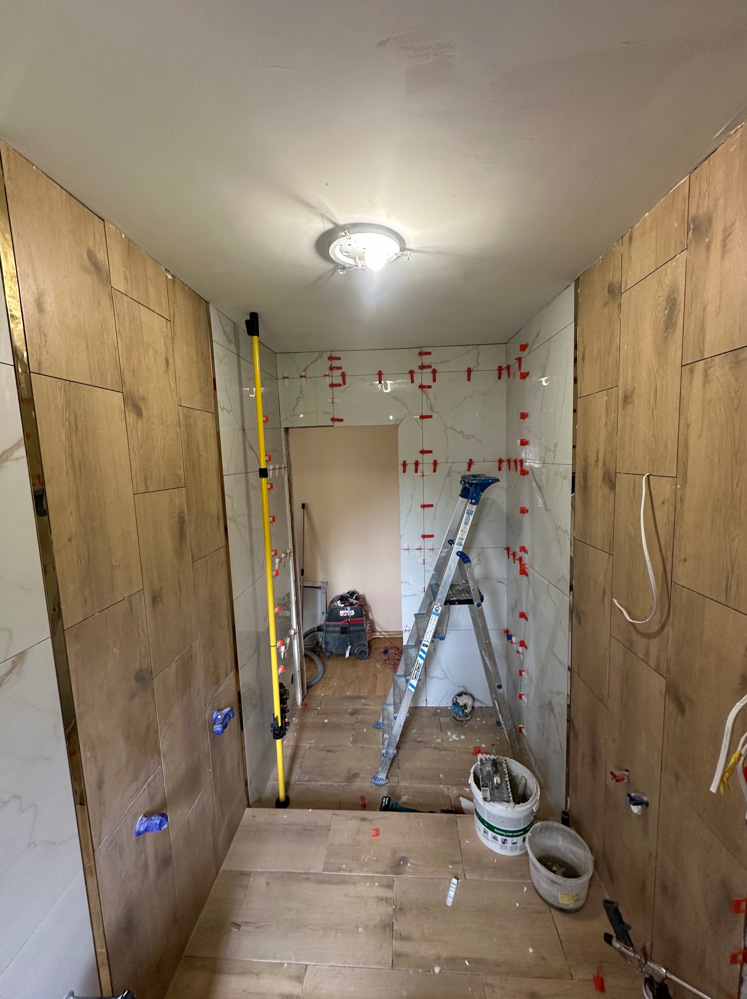 Wnętrze łazienki w trakcie prac glazurniczych: ułożone płytki imitujące drewno na ścianach i podłodze, poziomica, drabina i akcesoria glazurnicze.