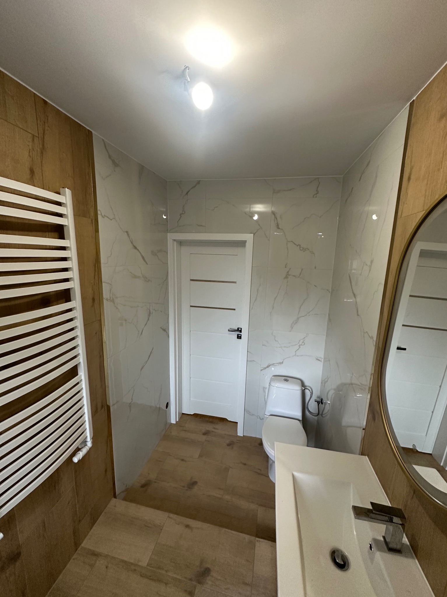 Nowoczesna łazienka po remoncie: białe i brązowe płytki imitujące drewno, grzejnik drabinkowy, umywalka z lustrem i toaleta. Jasne, przestronne wnętrze.