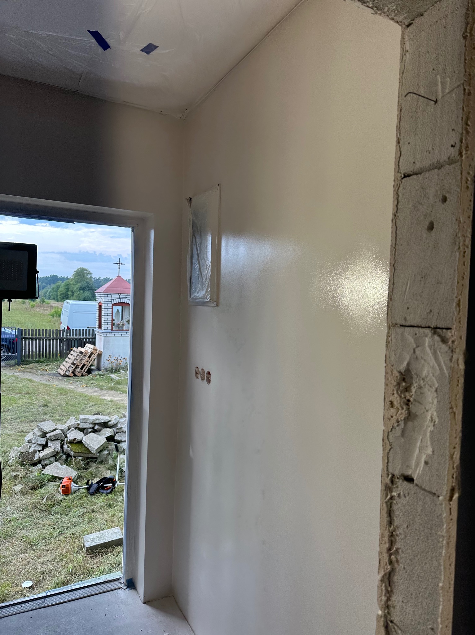 Świeżo pomalowana, gładka ściana w jasnym odcieniu wewnątrz budynku, widoczne zabezpieczenia folią malarską na suficie i gniazdkach. Surowy betonowy otwór drzwiowy.