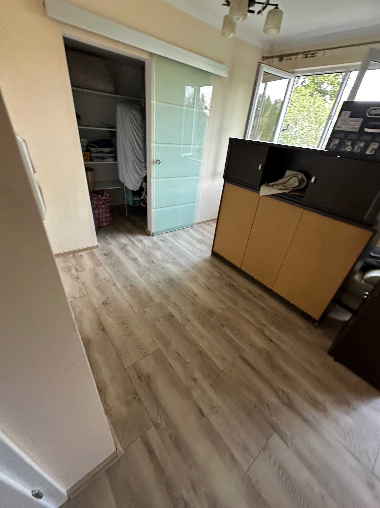 Jasne panele podłogowe w pokoju z przesuwnymi drzwiami do garderoby i komodą. Widok na okno z zielenią w tle. Ukończone układanie paneli.