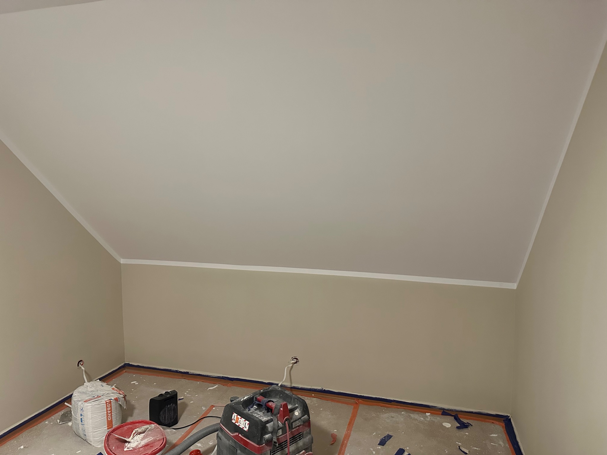 Pokój na poddaszu po szpachlowaniu i malowaniu, przygotowany do dalszych prac wykończeniowych. Widoczny odkurzacz przemysłowy i worki z materiałami budowlanymi.
