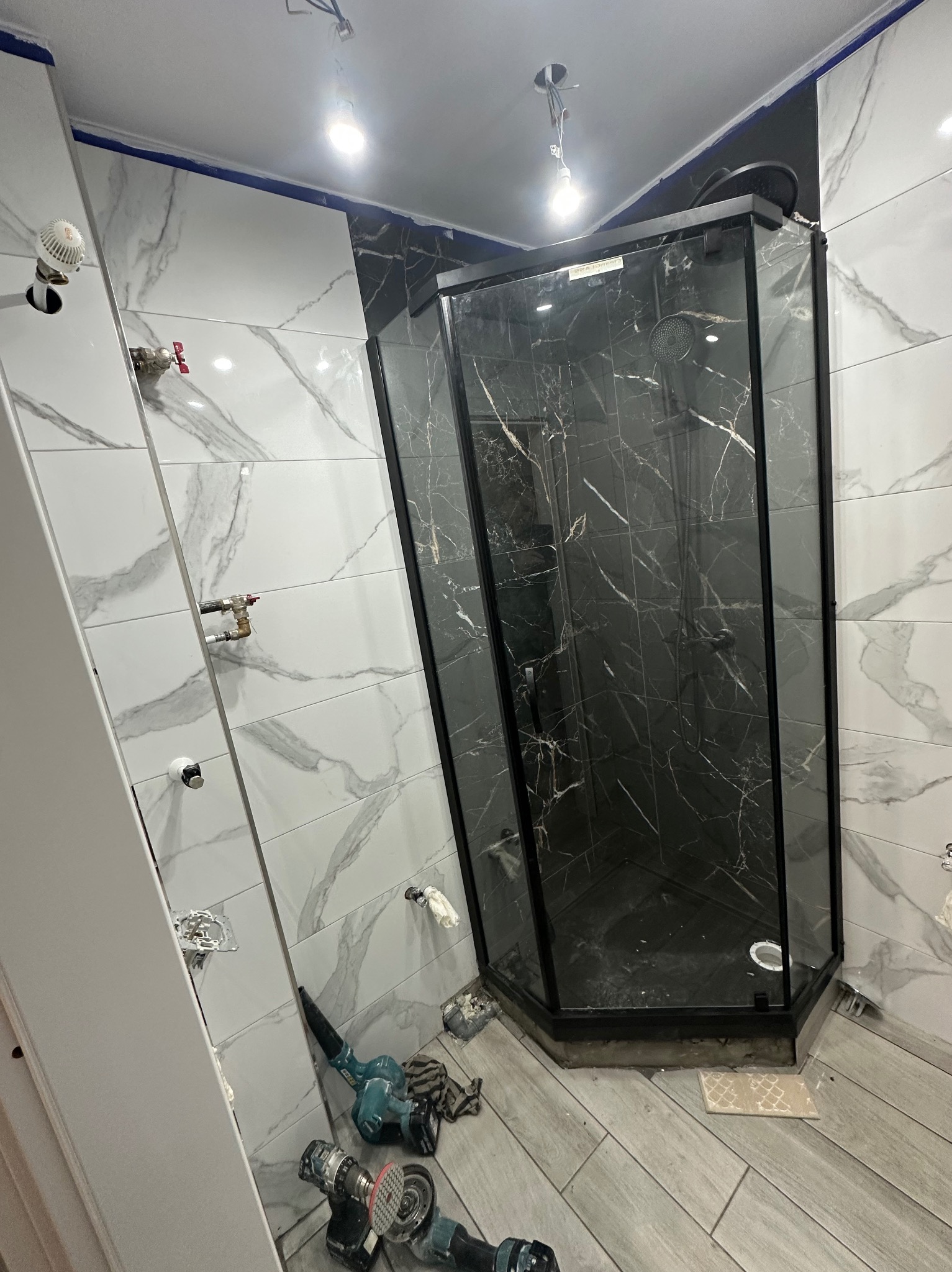 Nowoczesna kabina prysznicowa z czarnymi marmurkowymi płytkami w trakcie montażu. Widoczne narzędzia glazurnicze i białe płytki imitujące marmur na ścianach.