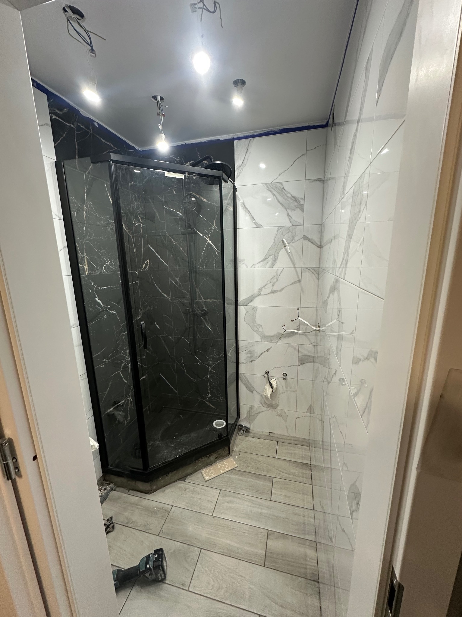 Nowoczesna kabina prysznicowa z czarną ramą i marmurowym wzorem płytek, szare płytki podłogowe, widoczne niedokończone prace elektryczne.