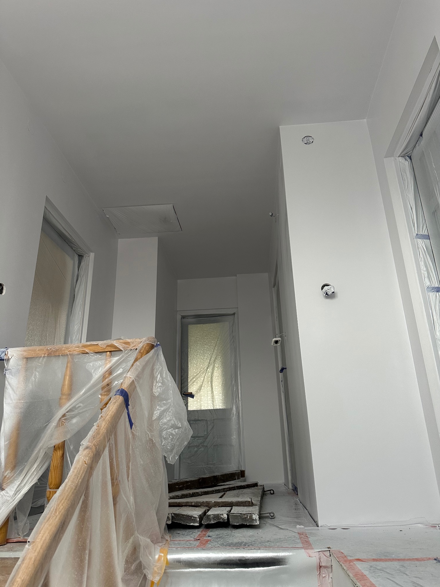 Białe, świeżo pomalowane wnętrze w trakcie prac wykończeniowych. Zabezpieczone folią okna i schody. Widoczne ślady po taśmie malarskiej na podłodze.