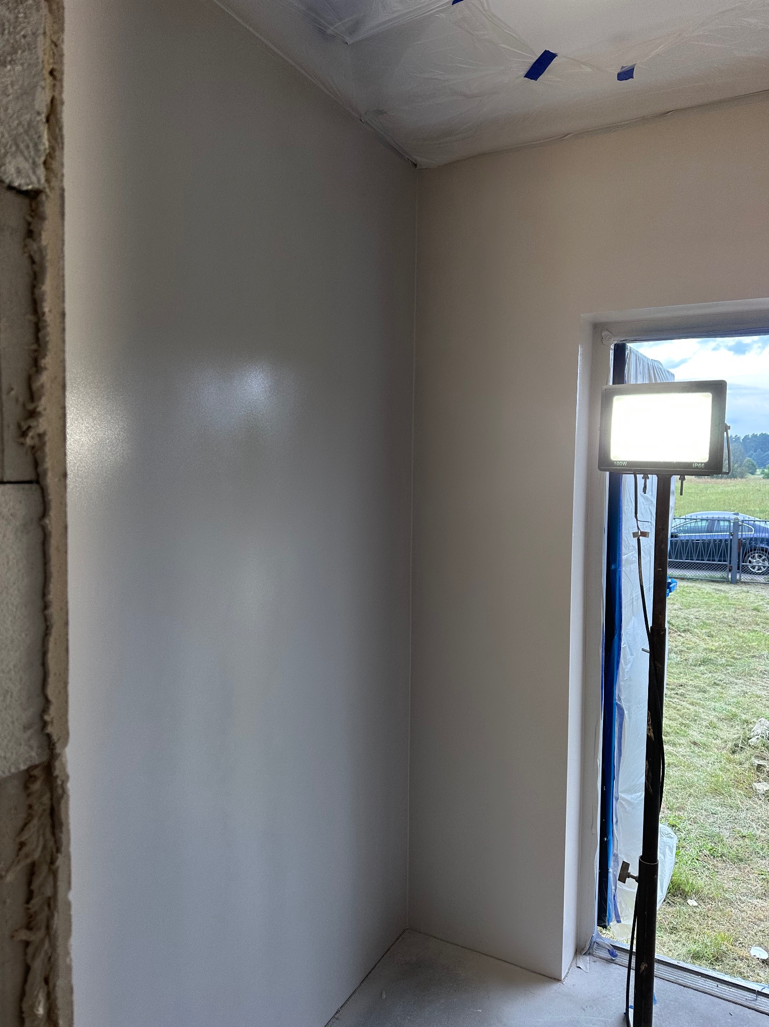 Gładka, świeżo pomalowana ściana w jasnym odcieniu szarości, oświetlona lampą budowlaną. Fragment surowego muru po lewej stronie. Okno z widokiem na zewnątrz.
