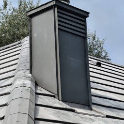 KAMILBUD - Szary komin wentylacyjny z metalową obudową i ukośnymi żaluzjami na dachu pokrytym szarą dachówką, widok z dołu, fragment drzewa w tle.