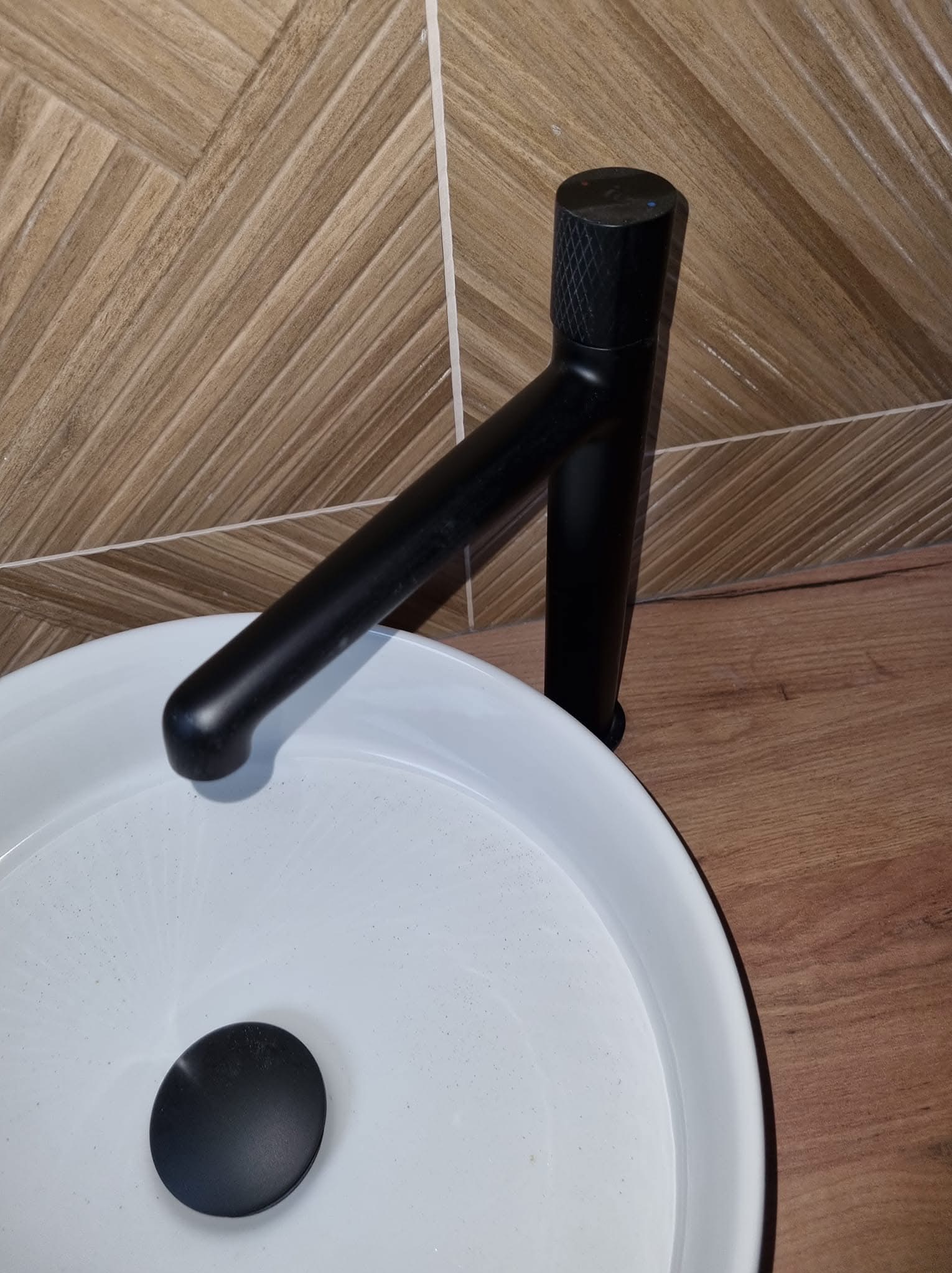 Biała umywalka nablatowa z czarną, matową baterią na tle drewnopodobnych płytek ściennych i blatu. Nowoczesny design.