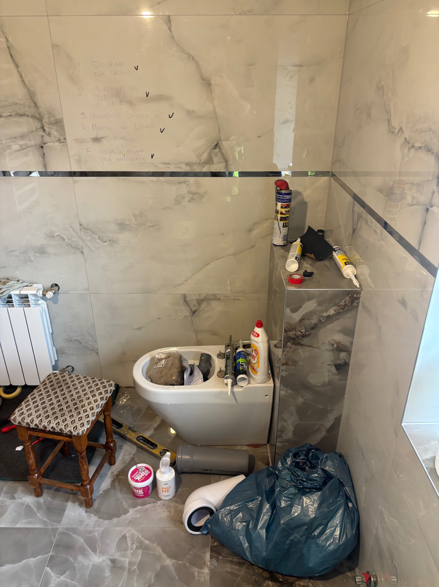 Remont łazienki w toku: białe płytki imitujące marmur, bidet, grzejnik i narzędzia. Na ścianie notatki z zadaniami. Widoczny etap prac wykończeniowych.