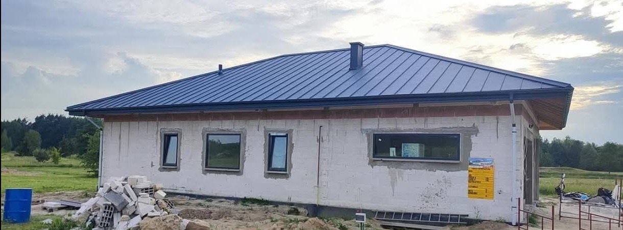 Nowoczesny dom w trakcie budowy z ciemnym dachem z blachy na tle zieleni. Widoczne okna i materiały budowlane.