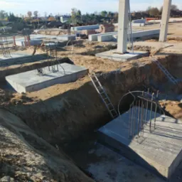 Wykopy pod fundamenty z częściowo wylanym betonem i wystającymi prętami zbrojeniowymi, drabiny w wykopach, przygotowane betonowe słupy w tle.
