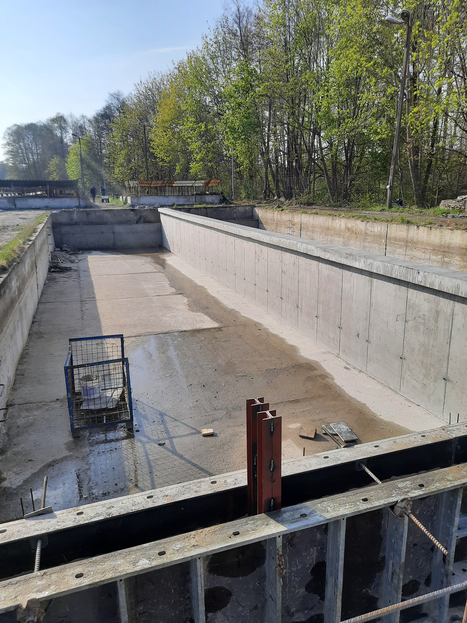 Surowy betonowy zbiornik w trakcie budowy, widoczne szalunki, zbrojenia i klatka z narzędziami, drzewa w tle.