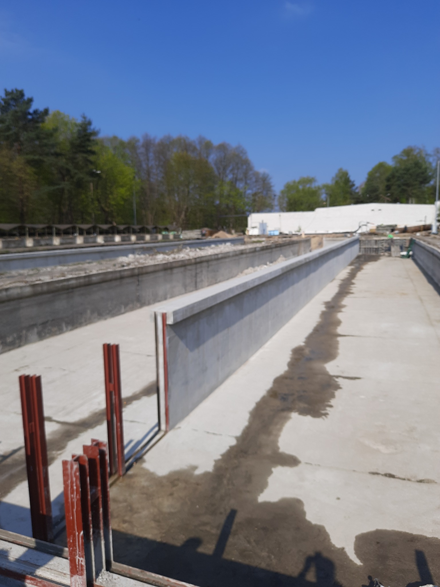 Betonowe koryto w trakcie budowy, z widocznymi czerwonymi elementami konstrukcyjnymi i śladami wilgoci na powierzchni betonu, w tle drzewa i błękitne niebo.