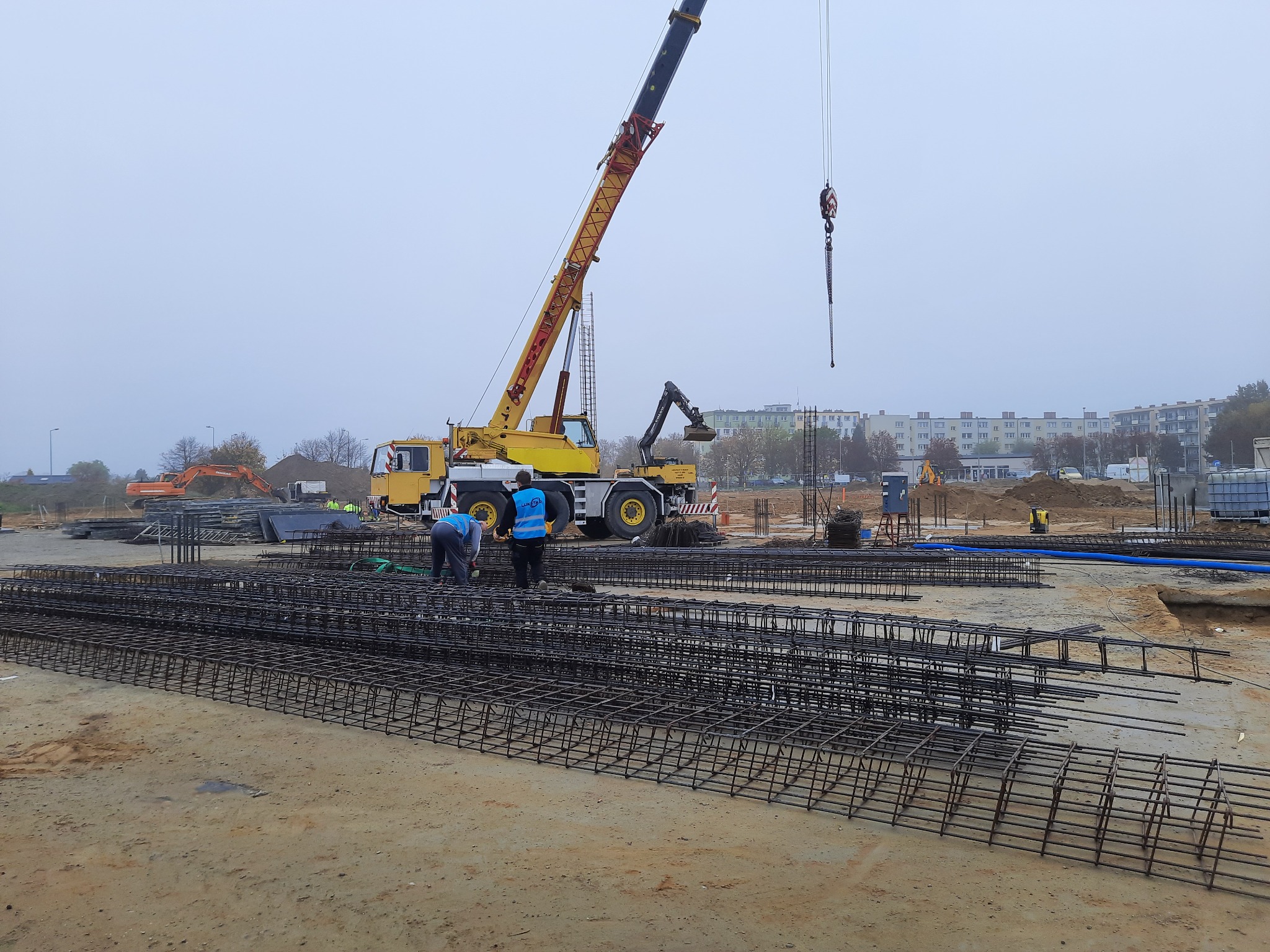 Zbrojenie fundamentów w postaci gotowych szkieletów z prętów stalowych na placu budowy, w tle żuraw budowlany i pracownicy w odblaskowych kamizelkach.