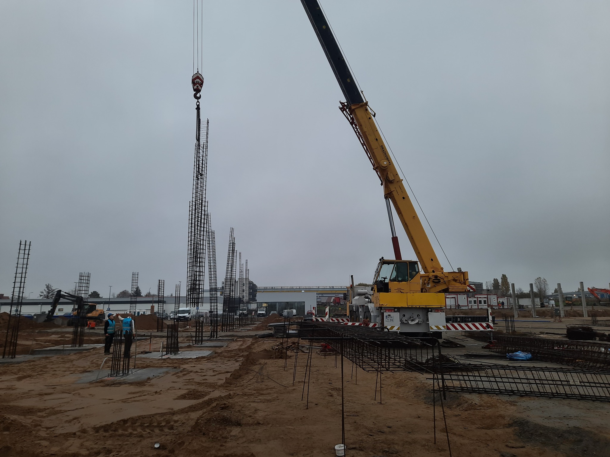 Żuraw podnosi zbrojenie słupa na placu budowy, widoczni pracownicy w odblaskowych kamizelkach, w tle inne elementy konstrukcyjne i pojazdy.