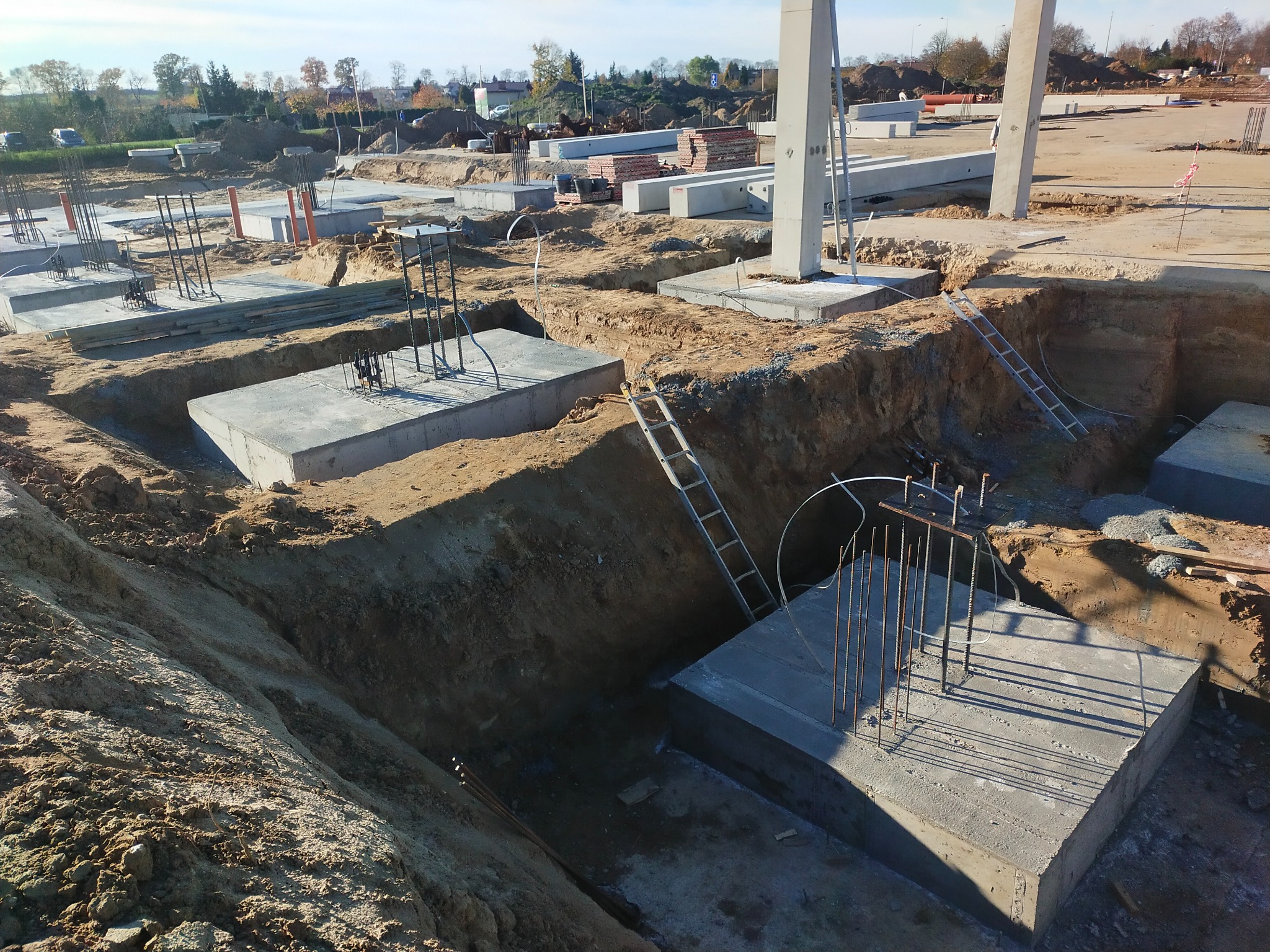 Wykopy pod fundamenty z częściowo wylanym betonem i wystającymi prętami zbrojeniowymi, drabiny w wykopach, przygotowane betonowe słupy w tle.