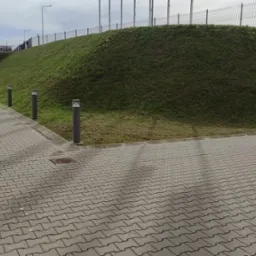 Uporządkowany teren zewnętrzny z kostki brukowej, przy budynku z panelami i trawiastym nasypem z lampami parkowymi, po świeżym skoszeniu trawy.