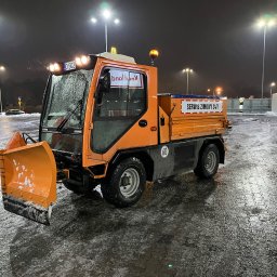 Tom & Jerry Garden - Pomarańczowy pojazd do odśnieżania marki Ladog z napisem 'Serwis Zimowy 24/7' na tle nocnego parkingu po opadach śniegu, oświetlonego przez wysokie latarnie.