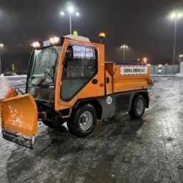 Pomarańczowy pojazd do odśnieżania marki Ladog z napisem 'Serwis Zimowy 24/7' na tle nocnego parkingu po opadach śniegu, oświetlonego przez wysokie latarnie.