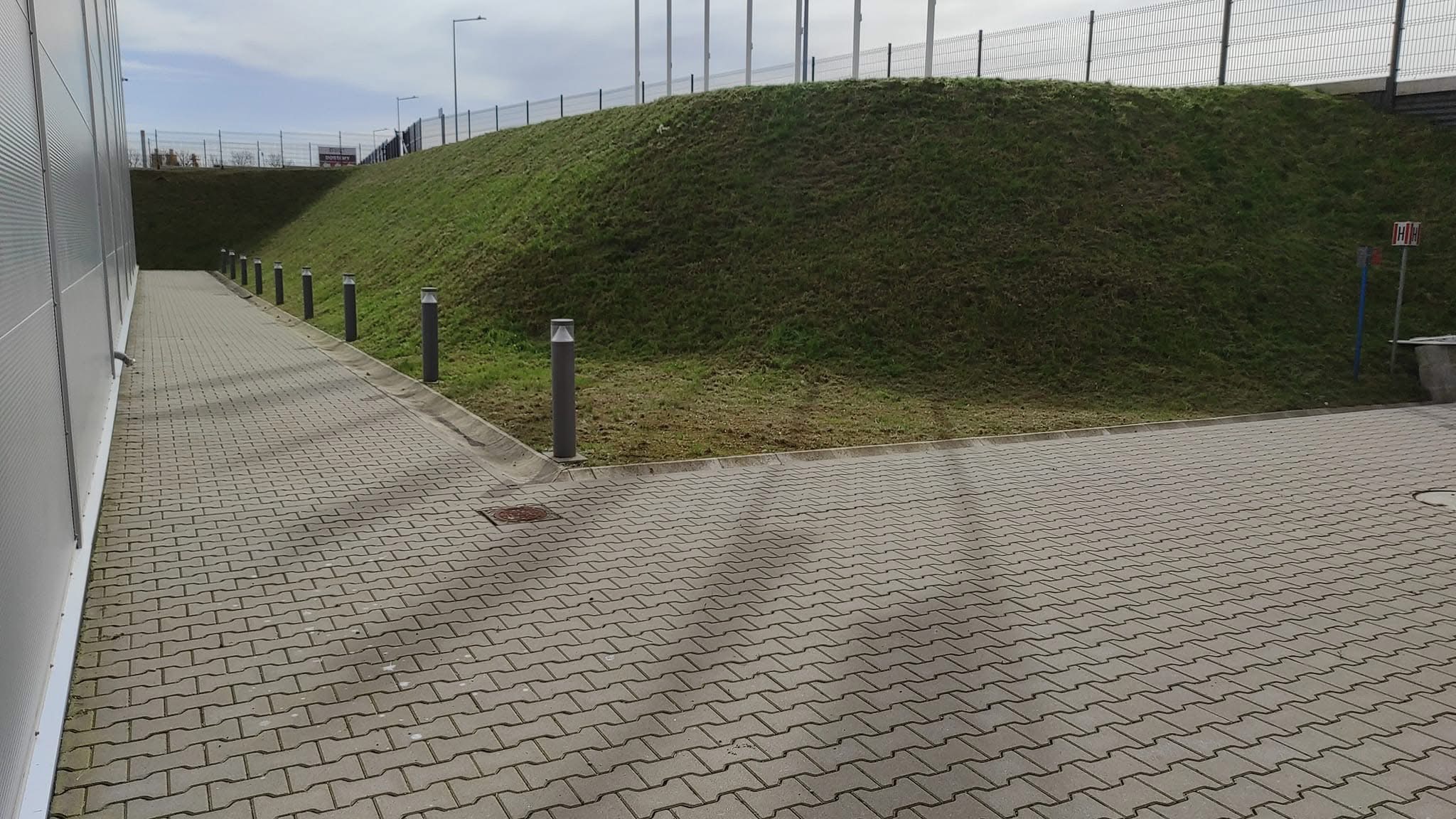 Uporządkowany teren zewnętrzny z kostki brukowej, przy budynku z panelami i trawiastym nasypem z lampami parkowymi, po świeżym skoszeniu trawy.