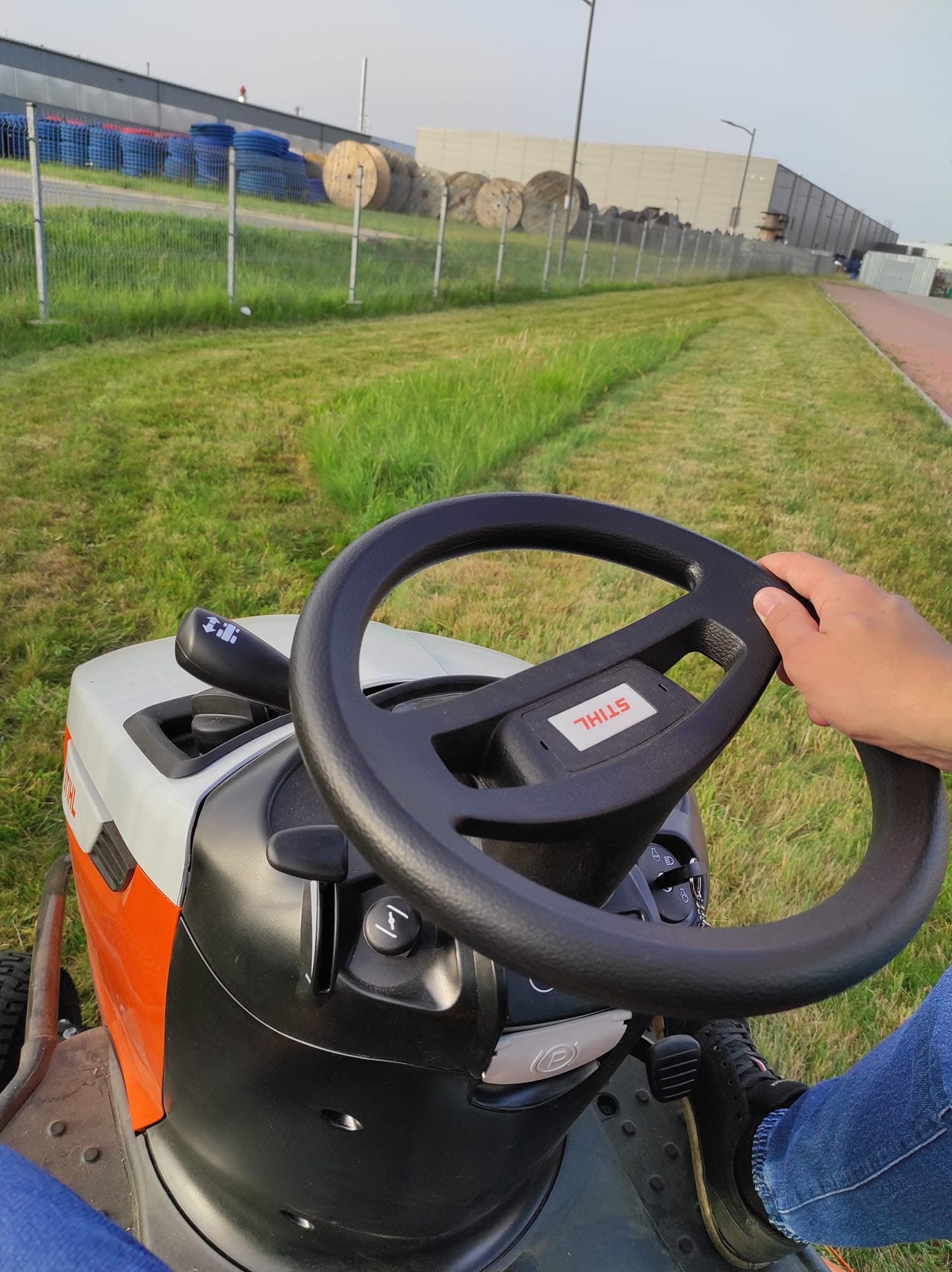 Operator kosiarki traktorka marki Stihl, w trakcie koszenia trawy na tle przemysłowym z widocznymi bębnami kablowymi i ogrodzeniem z siatki.