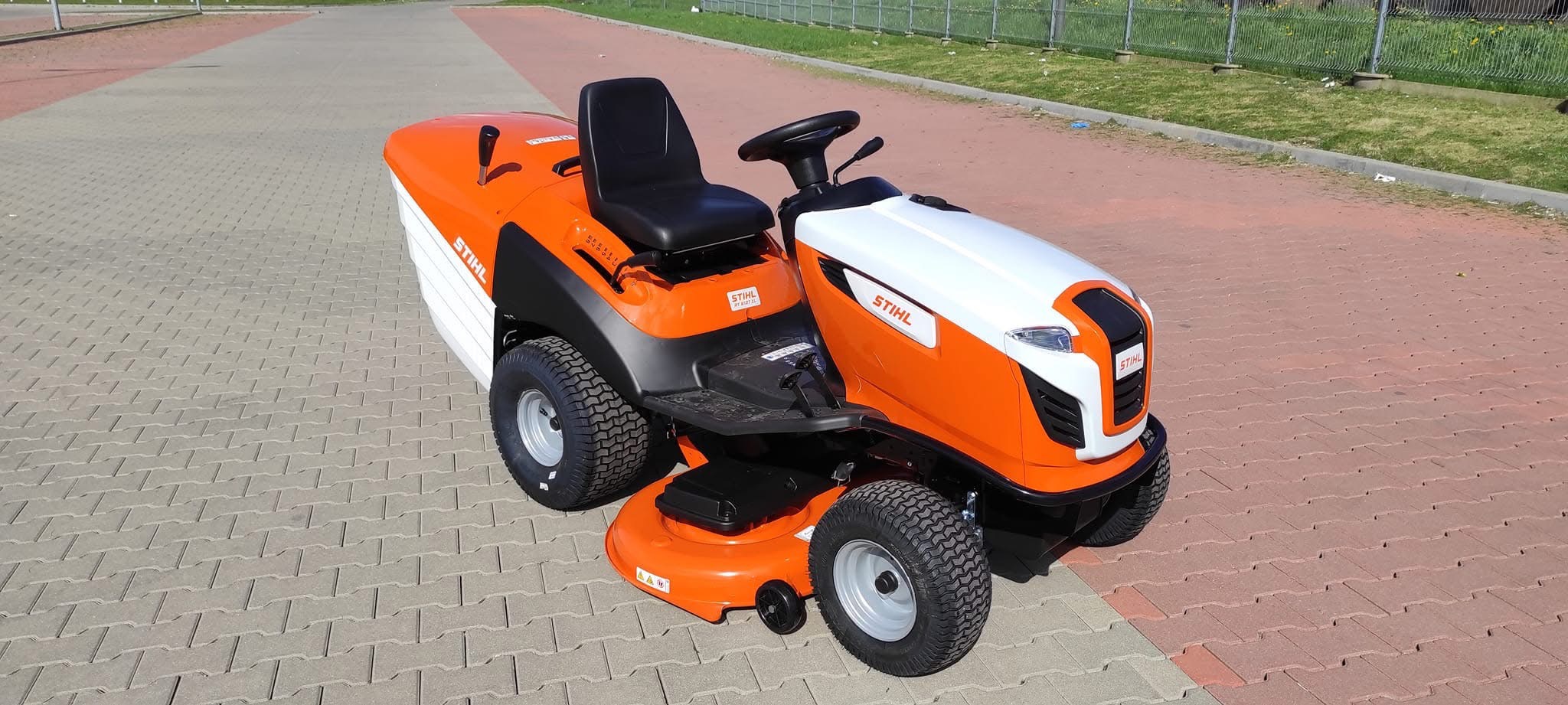 Pomarańczowo-biała kosiarka traktorowa marki Stihl, stojąca na kostce brukowej, z widocznym siedzeniem, kierownicą i logo producenta.