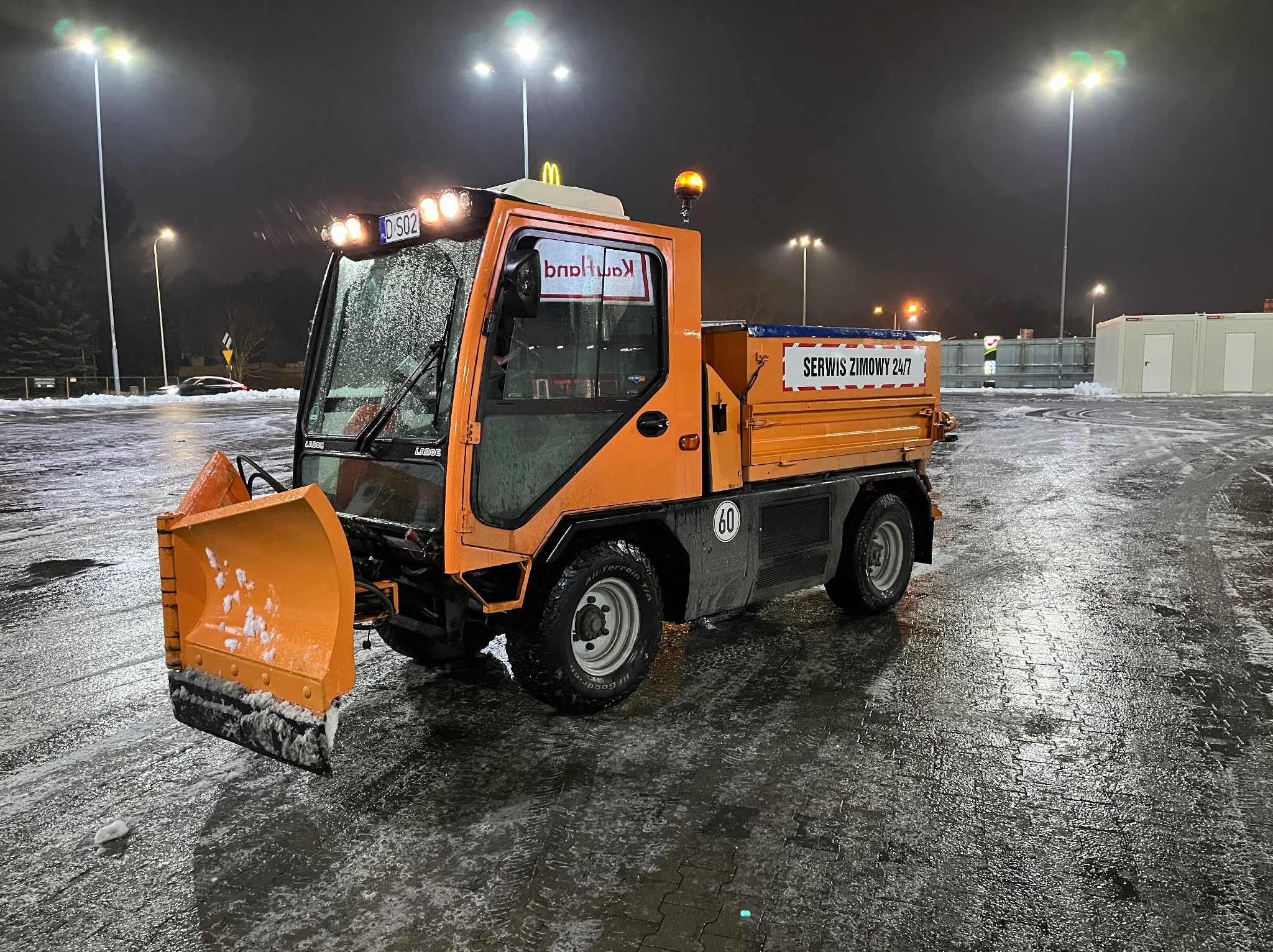 Pomarańczowy pojazd do odśnieżania marki Ladog z napisem 'Serwis Zimowy 24/7' na tle nocnego parkingu po opadach śniegu, oświetlonego przez wysokie latarnie.