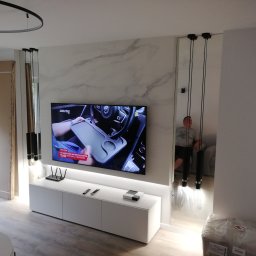 Promax ekskluzywne wnętrza - Salon z telewizorem na ścianie imitującej marmur, biała szafka RTV z dekoderem i routerem, lustrzane kolumny po bokach, czarne lampy wiszące, widoczne fragmenty niezakończonych prac wykończeniowych.