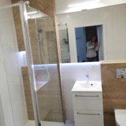 Promax ekskluzywne wnętrza - Nowoczesna łazienka po remoncie: kabina prysznicowa ze szklanymi drzwiami, biała umywalka z szafką, lustro z oświetleniem i drewniane akcenty na ścianach.