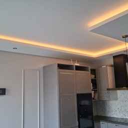 Promax ekskluzywne wnętrza - Nowoczesna kuchnia w szarościach z podświetlanym sufitem i złotymi lampami wiszącymi. Widoczny fragment ściany z uchwytem na TV i gniazdkami.