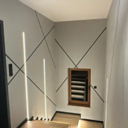 Nowoczesne wykończenie wnętrza: szare ściany z czarnymi liniami i pionowe LED-owe oświetlenie. Drewniane schody z podświetleniem.