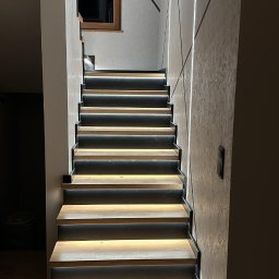 Promax ekskluzywne wnętrza - Nowoczesne schody z podświetleniem LED, drewniane stopnie z czarnymi obramowaniami, ściana z dekoracyjną fakturą i geometrycznymi wzorami, okno w tle.