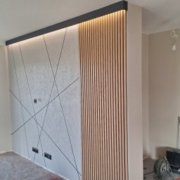 Promax ekskluzywne wnętrza - Nowoczesna ściana z dekoracyjną strukturą i drewnianymi lamelami podświetlonymi od góry. Widoczne gniazdka elektryczne i fragment okna. Podłoga zabezpieczona folią.