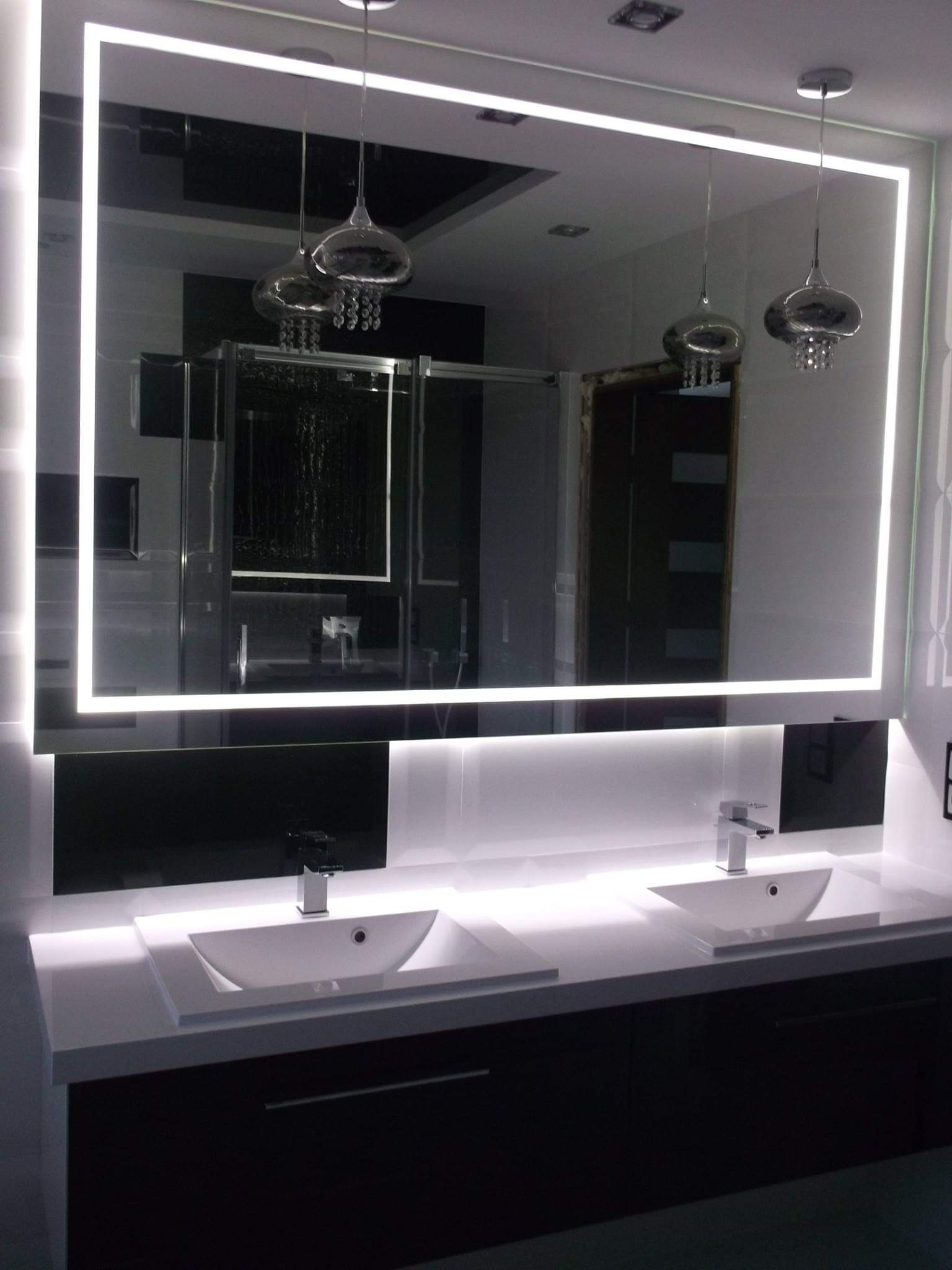 Nowoczesna łazienka z podświetlanym lustrem, dwie białe umywalki nablatowe na czarnym blacie, wiszące lampy dekoracyjne, czarno-białe płytki na ścianie.