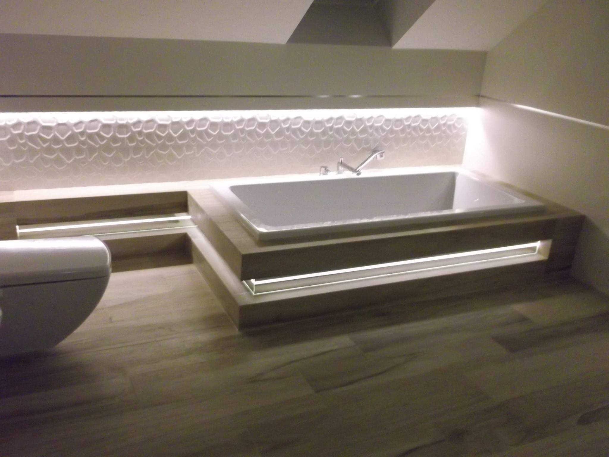 Nowoczesna łazienka z podświetlaną wanną w zabudowie, dekoracyjna mozaika na ścianie, drewnopodobne płytki na podłodze, oświetlenie LED.