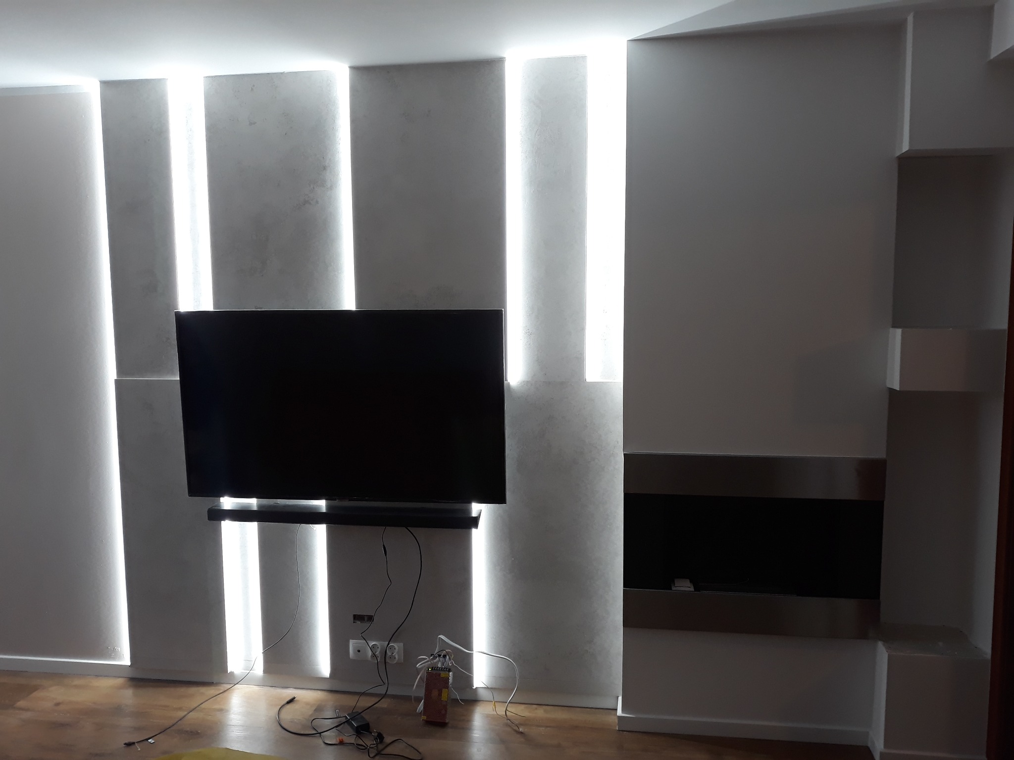 Nowoczesny salon z podświetlaną ścianą z szarego tynku strukturalnego, wiszący telewizor z widocznymi kablami i wbudowany kominek ze stalowym obramowaniem.