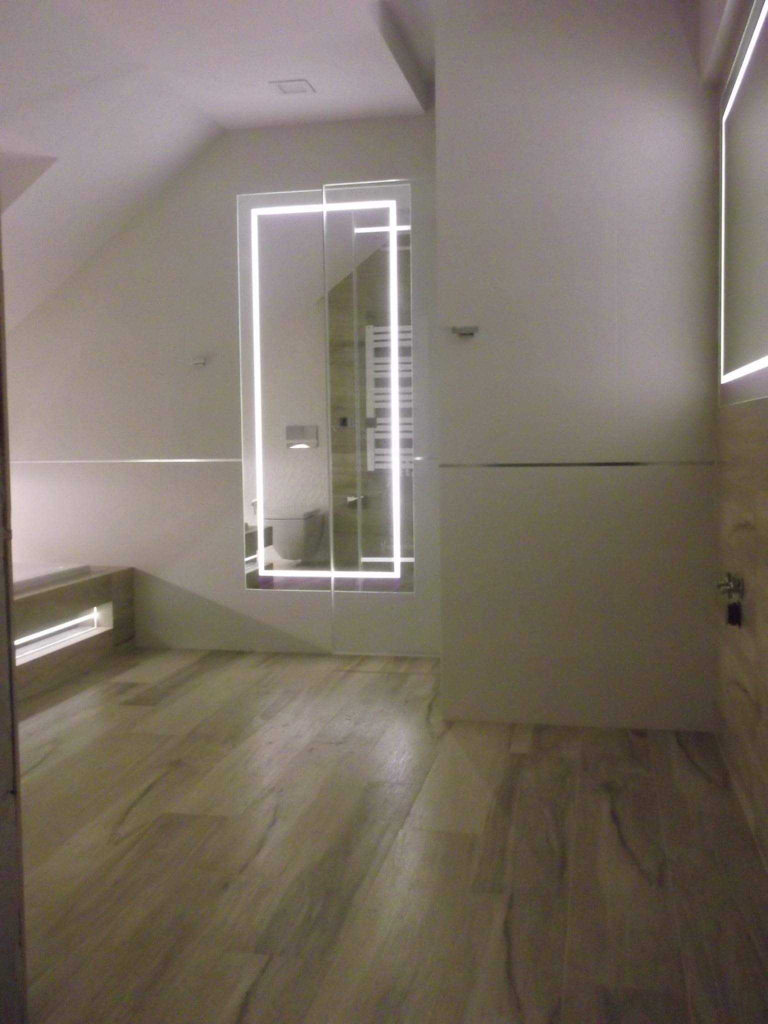 Wykończona łazienka na poddaszu z drewnopodobną podłogą, podświetlanym lustrem i wanną w zabudowie.