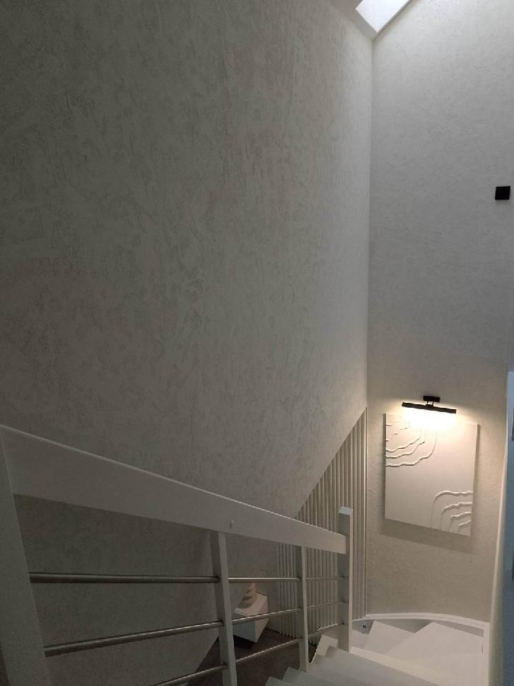 Klatka schodowa z wykończonymi ścianami w jasnych odcieniach, z elementami dekoracyjnymi i oświetleniem. Widoczne schody z białą balustradą i fragment okna dachowego.