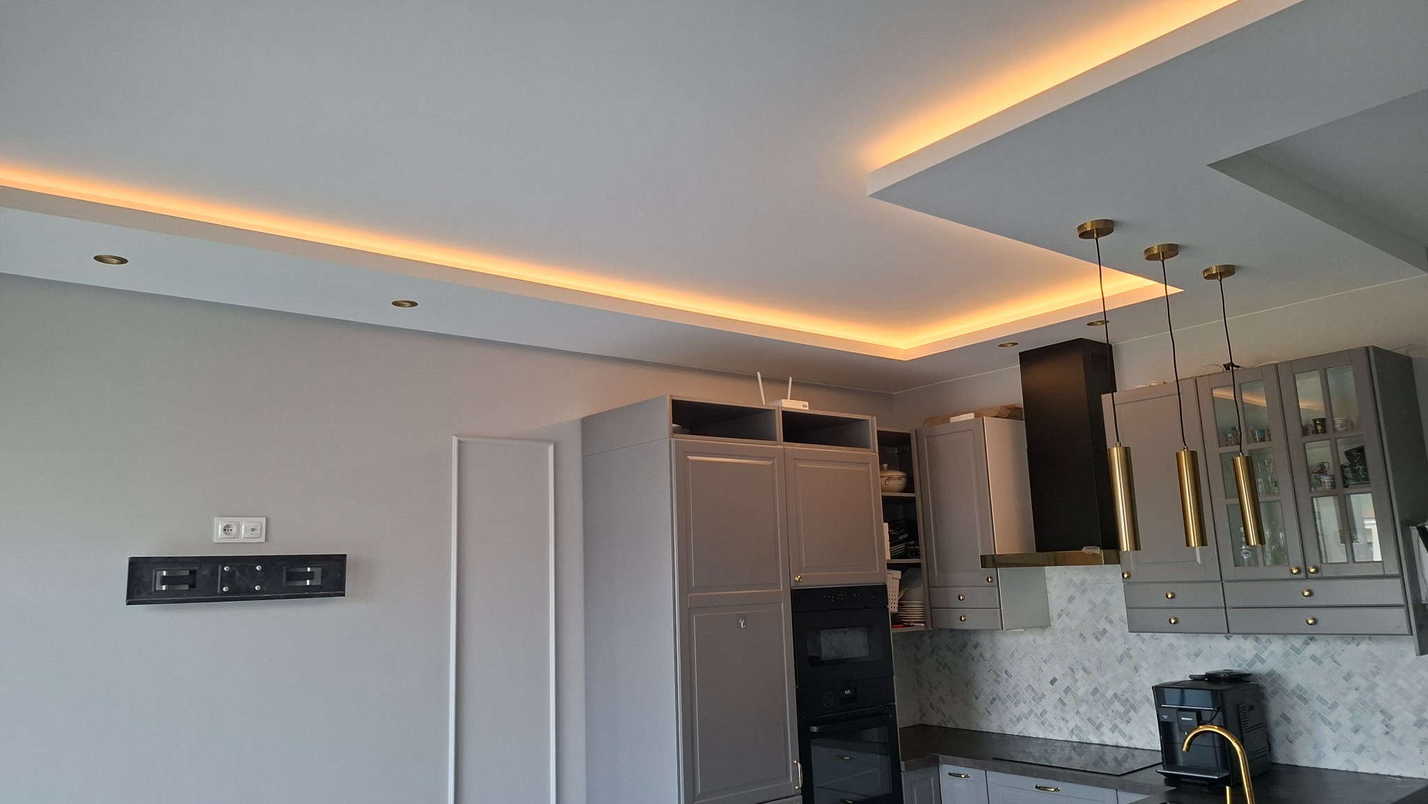 Nowoczesna kuchnia w szarościach z podświetlanym sufitem i złotymi lampami wiszącymi. Widoczny fragment ściany z uchwytem na TV i gniazdkami.