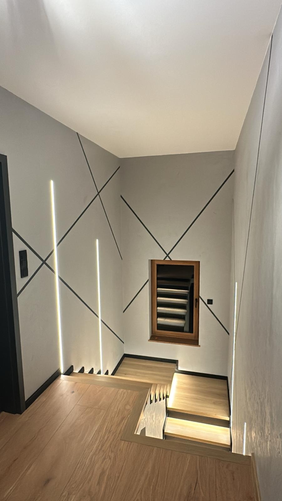 Nowoczesne wykończenie wnętrza: szare ściany z czarnymi liniami i pionowe LED-owe oświetlenie. Drewniane schody z podświetleniem.