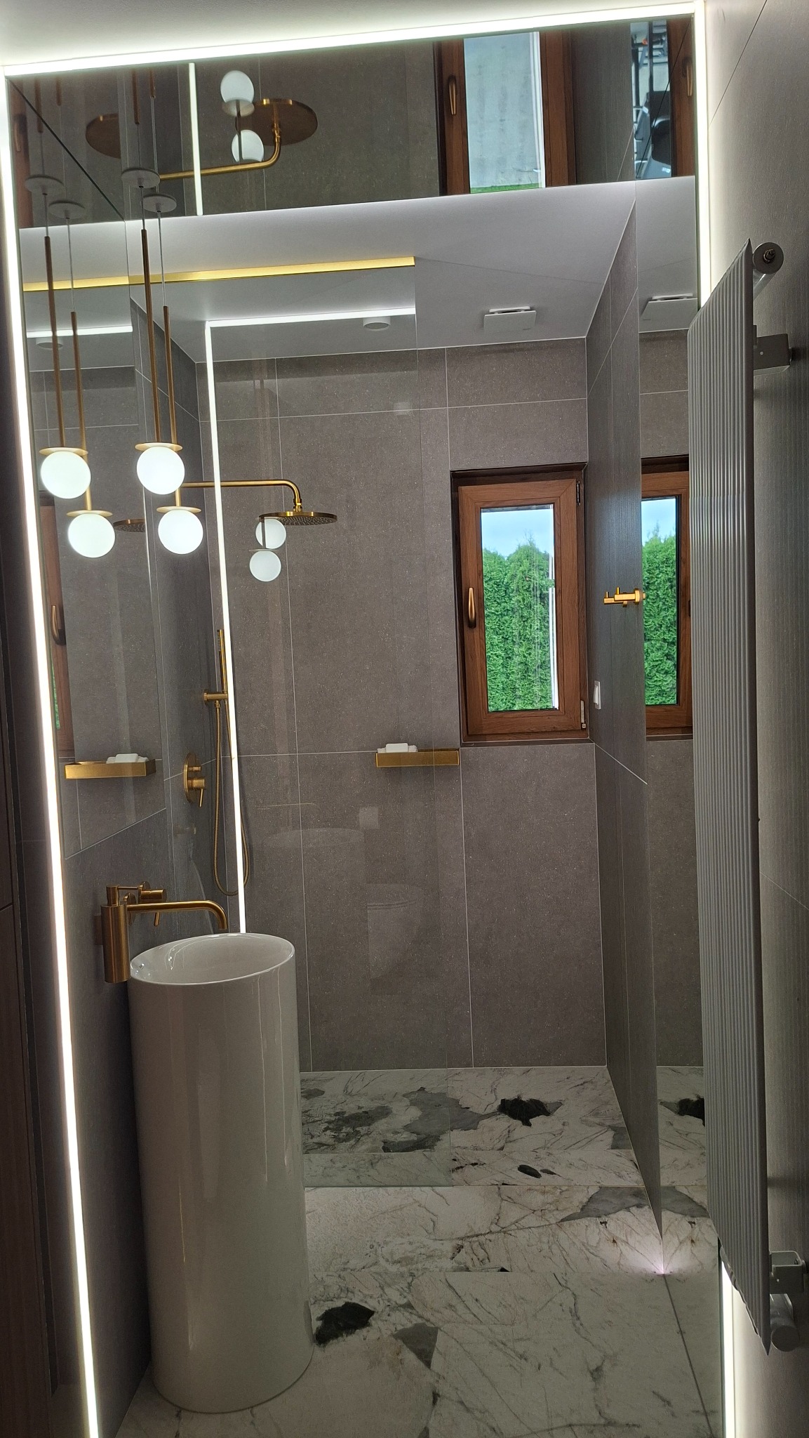 Nowoczesna łazienka z szarą płytką, marmurową podłogą i złotymi akcentami: armatura, oświetlenie, półki. Umywalka wolnostojąca, okno z widokiem na zieleń.