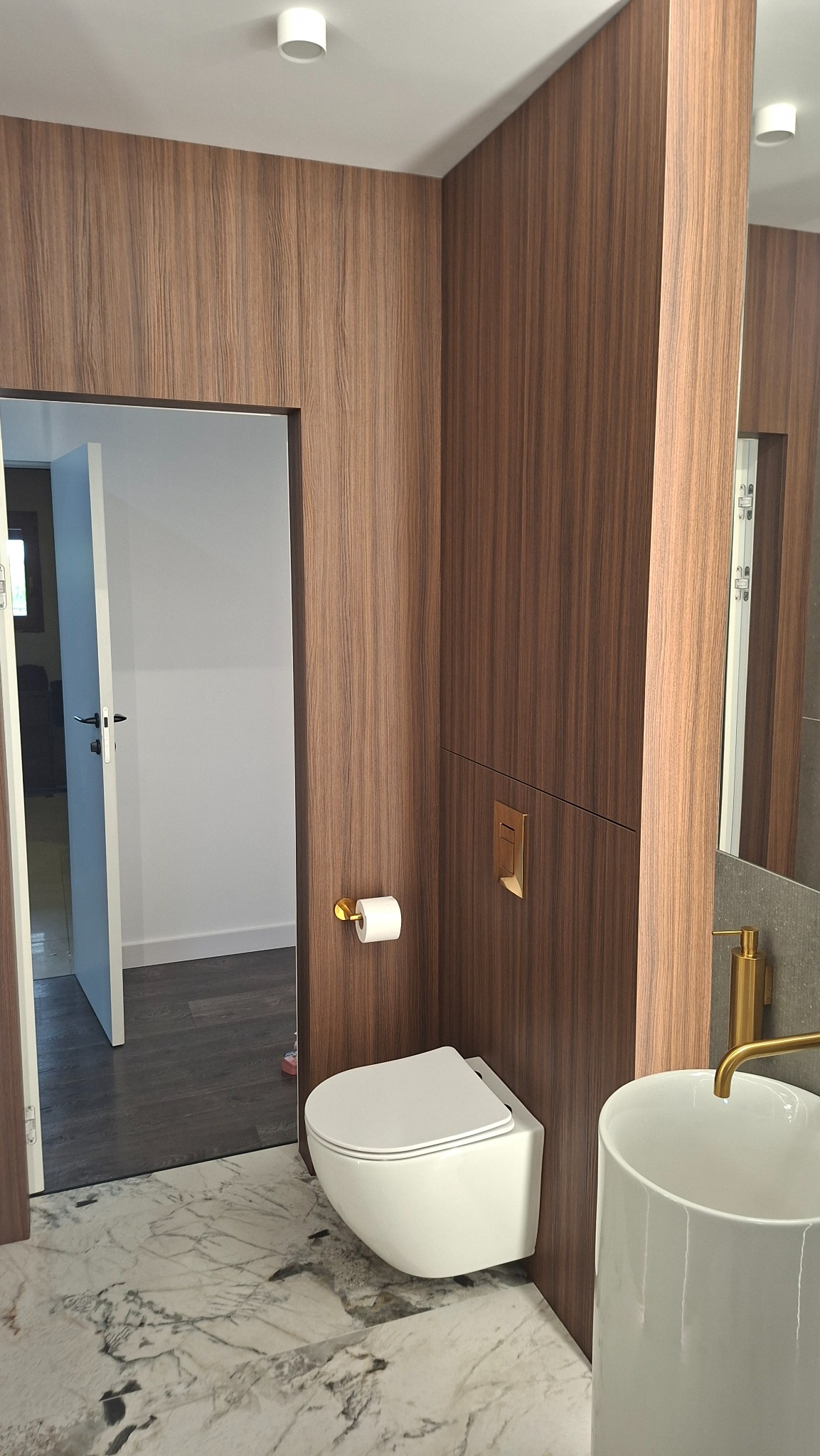 Nowoczesna łazienka z drewnianą okładziną ścienną, wiszącą toaletą i marmurową podłogą. Złote akcenty armatury dodają elegancji. Widok na otwarte drzwi.