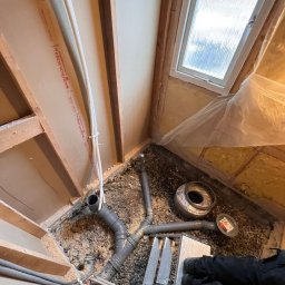 AdamBygg - Fragment łazienki w trakcie prac: widoczne rury kanalizacyjne na podłożu z drobnych kamieni, częściowo obudowane ścianami z płyt gipsowo-kartonowych i drewnianą konstrukcją, małe okno w tle.