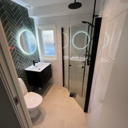 AdamBygg - Nowoczesna łazienka z czarnymi akcentami: kabina prysznicowa ze szklanymi drzwiami, umywalka na szafce, toaleta i podświetlane okrągłe lustro na ścianie z ciemnej, ułożonej w jodełkę glazury.