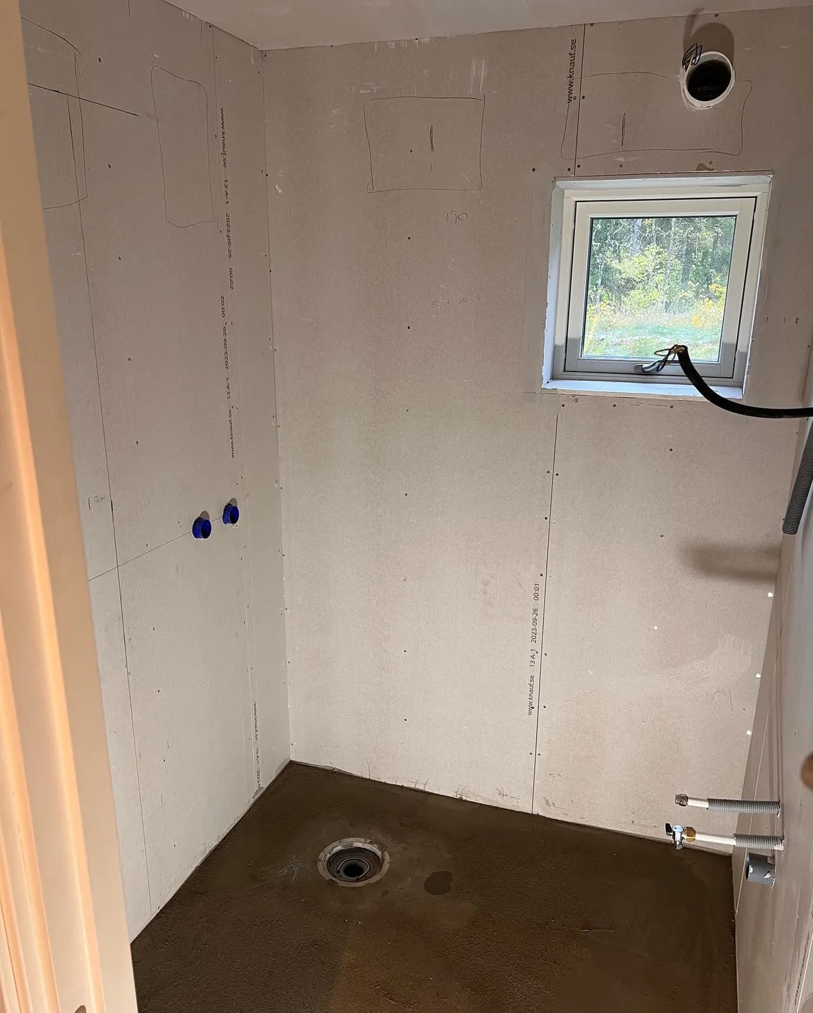 Surowa łazienka w trakcie przygotowania do wykończenia: widoczne płyty karton-gips na ścianach, wylewka na podłodze z odpływem oraz wystające rury instalacji wodnej i okno z widokiem na zieleń.
