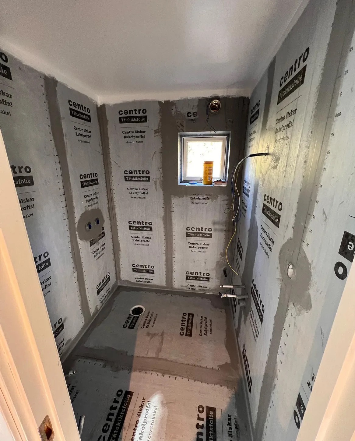 Łazienka w trakcie remontu: ściany i podłoga pokryte folią uszczelniającą z logo Centro, widoczne rury instalacyjne i okno z żółtym pojemnikiem na parapecie.